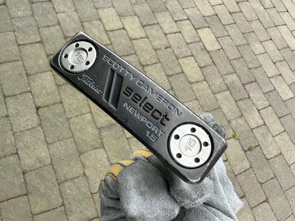 Close-up of a black Scotty Cameron Select Newport 15 putter head with weights, held by a gloved hand over brick pavement.