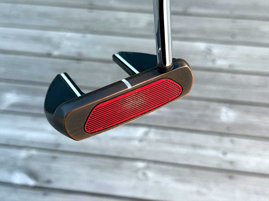 Close-up of a mallet-style golf putter head with a red grooved face insert and a chrome shaft, set against a wooden deck background.
