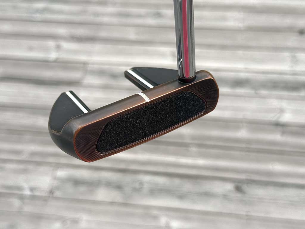 Copper-finished golf putter head with a black face insert and white alignment line, mounted on a shaft against a blurred wooden deck background.